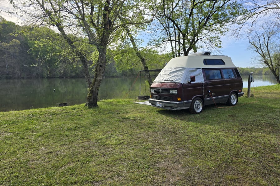 Relaxing by the river: a maroon camper van, hooked up at a scenic spot, perfect for your next RV rental trip.