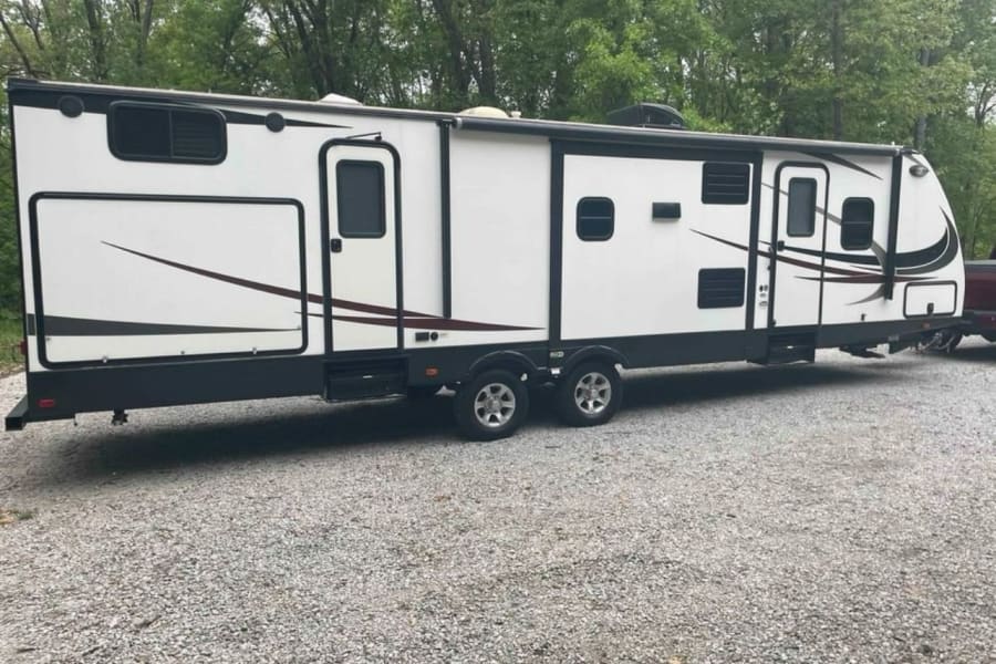 Side view of a long white & black travel trailer RV rental with slide-outs, parked on a gravel lot.