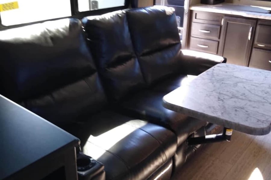 Bright rv rental interior: black reclining sofa, marble table, and kitchen area. Cozy, spacious, ready for adventure.