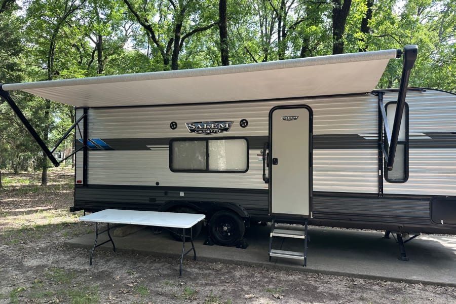 Modern travel trailer RV rental with awning extended, parked on a concrete pad at a wooded campsite, ready for your trip.