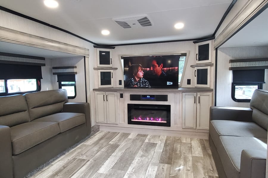 Modern RV rental living space featuring comfy sofas, a large TV with an electric fireplace, and wood-look flooring.