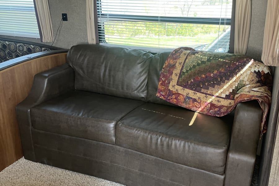 Relax in this RV rental's cozy dark faux leather sofa with a colorful quilt, window blinds, and wood cabinets.