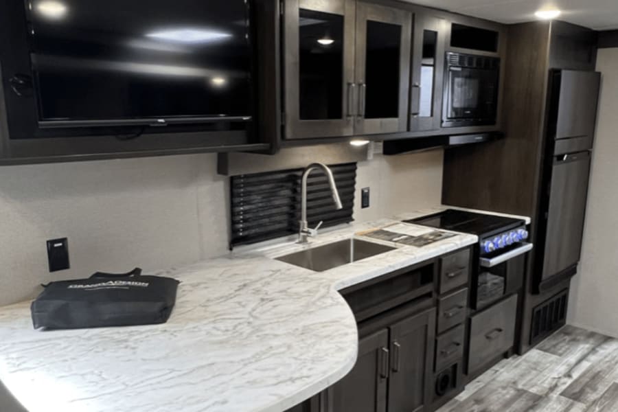 Modern RV rental interior features a full kitchen with sink, stove, microwave, fridge, and TV. Dark cabinets & marble counters.