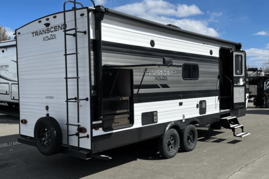 White and grey travel trailer, an RV rental. Features: rear ladder, extended awning, entry steps, and outdoor kitchen.