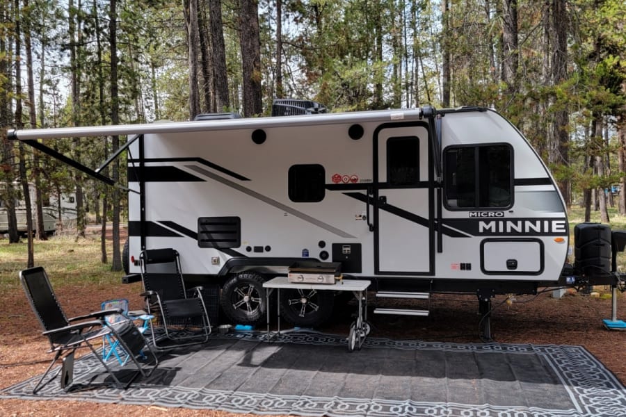 This Micro Minnie RV rental is perfectly set up at a wooded campsite with an extended awning, chairs, grill, and outdoor mat.