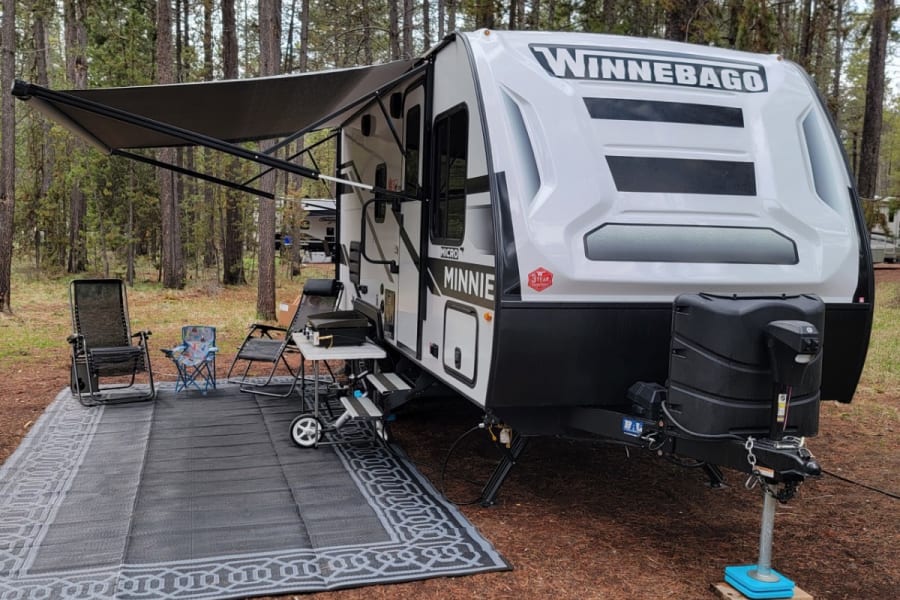 Winnebago Minnie Micro RV rental at a forest campsite with awning, chairs, and outdoor rug for comfortable living.