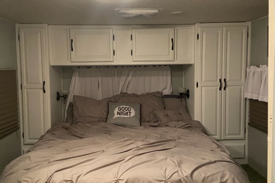 This cozy RV rental bedroom features a bed with soft taupe bedding, white storage cabinets, and overhead lighting.