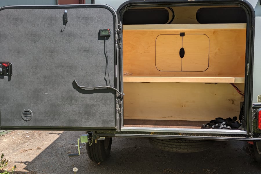 Open rear galley of an RV rental camper, revealing plywood shelves, storage, and a power monitor on the door.