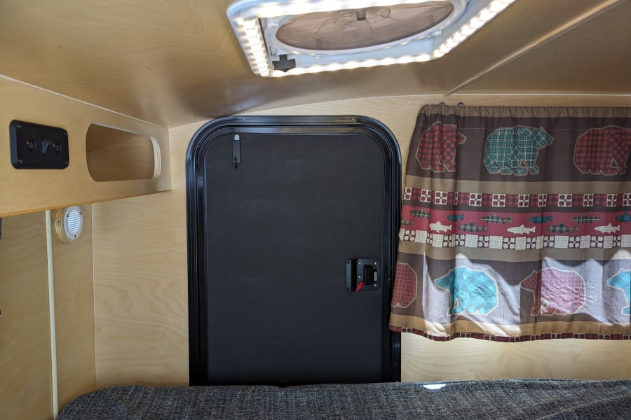 View inside a compact RV rental: wood-paneled interior, black door, ceiling fan/light, bear/fish curtain, and textured bed.