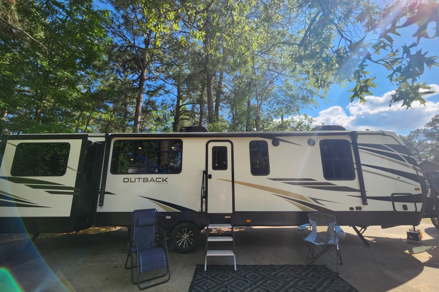 A large white and black Outback RV rental fully set up with outdoor chairs and a mat at a sunny campsite.