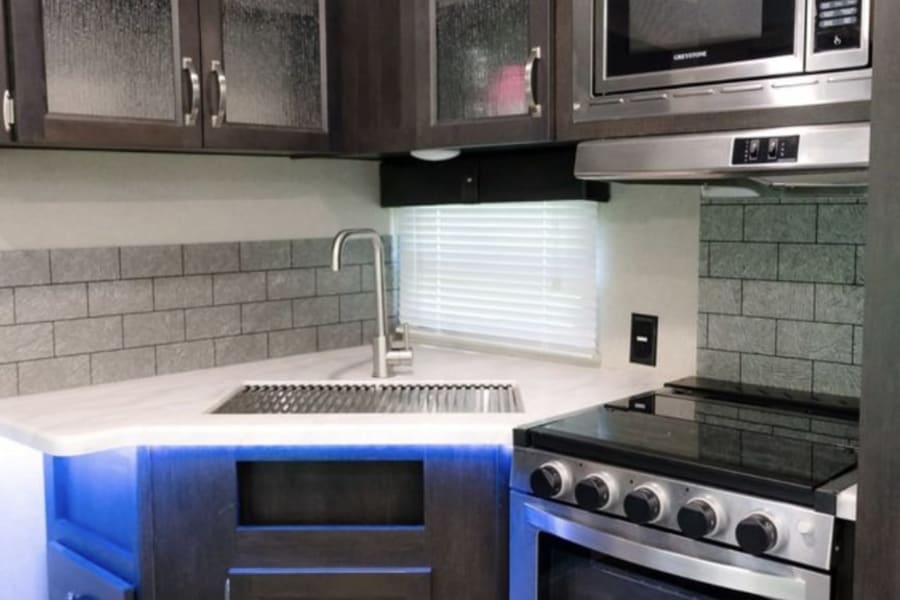 Modern RV rental kitchen featuring dark cabinetry, stainless steel microwave/stove, sink, and cool blue under-cabinet lighting.
