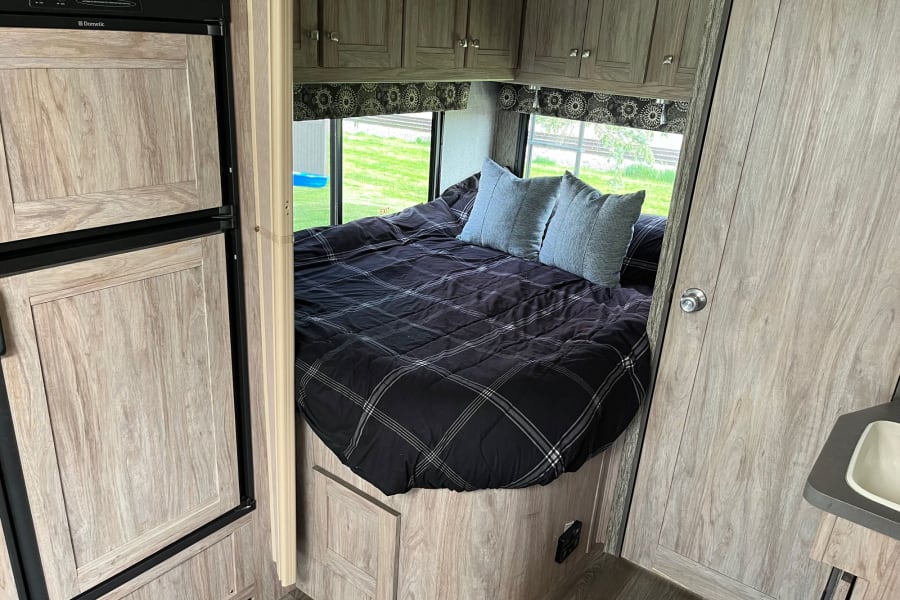 Interior of an RV rental: a cozy bedroom with a large bed, plaid comforter, and windows offering scenic views. Storage cabinets visible.