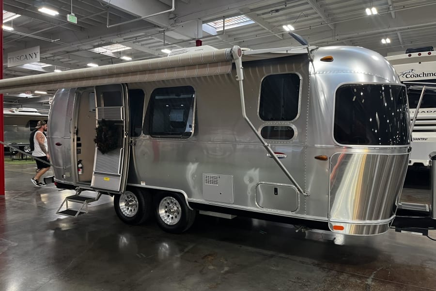 Shiny silver Airstream travel trailer with awning extended, perfect for your next RV rental. Displayed in an indoor showroom.