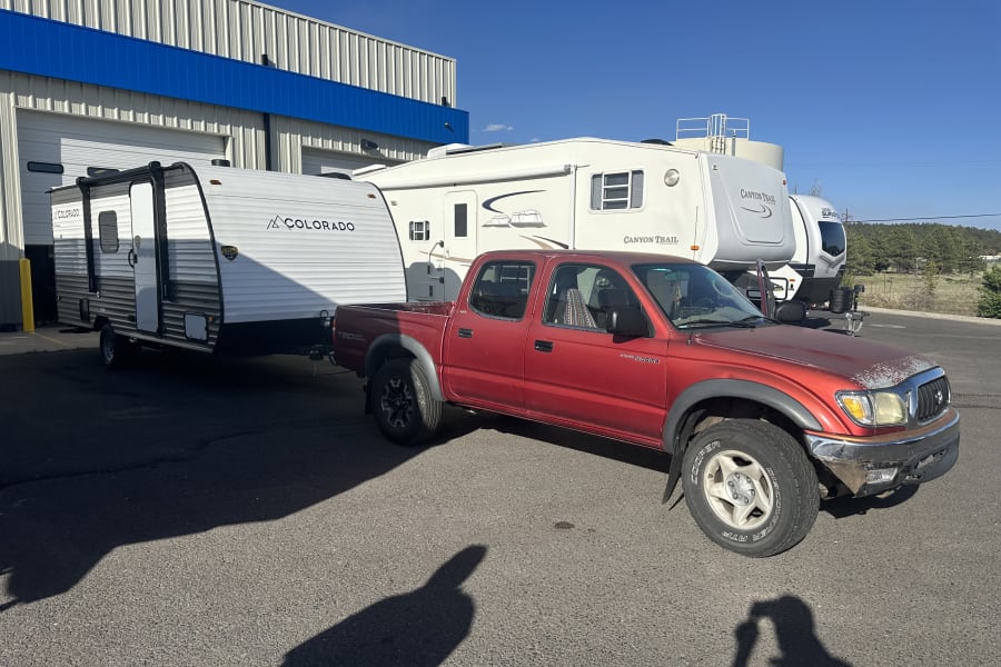 A red pickup truck is hitched to a white Colorado travel trailer, potentially an RV rental, with other RVs in a lot.