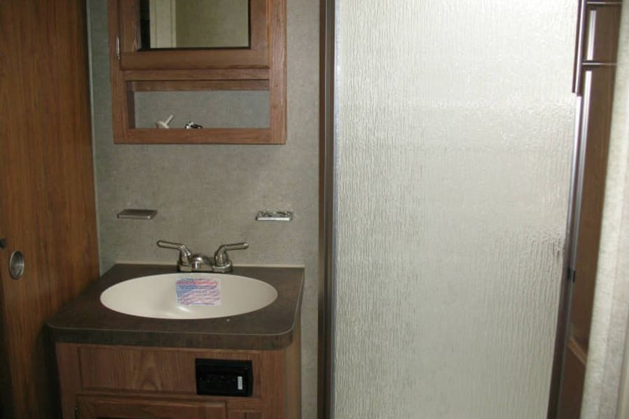 Inside an RV rental: a compact bathroom with a wood vanity, sink, mirror cabinet, and textured glass shower door.