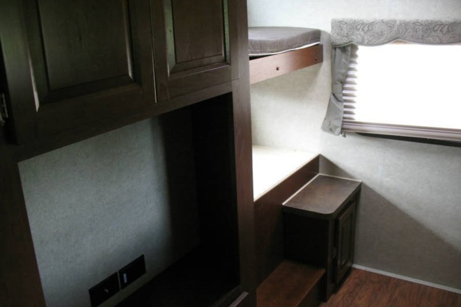 Experience comfort in this **rv rental**! Dark wood cabinets, a cozy bunk bed, and a window with blinds make a snug camper interior.