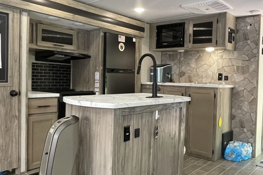 Inside an RV rental: a bright kitchen with a central island, light wood cabinetry, black accents, and a stone feature wall.