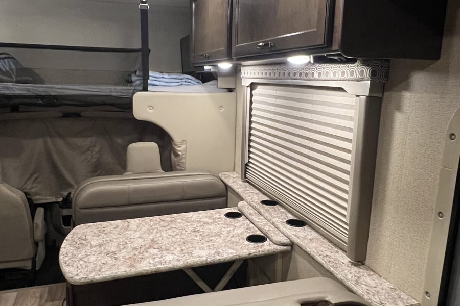 RV rental interior: dinette with table and cup holders, overhead cabinets, and bunk bed with safety net visible.