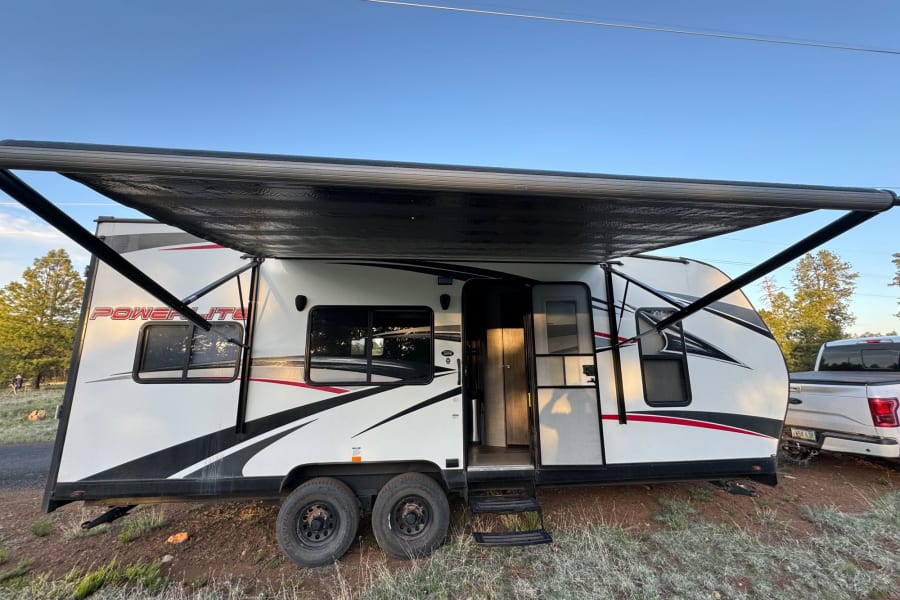 A white Power Lite toy hauler RV rental with an extended black awning, parked outdoors in a natural setting.