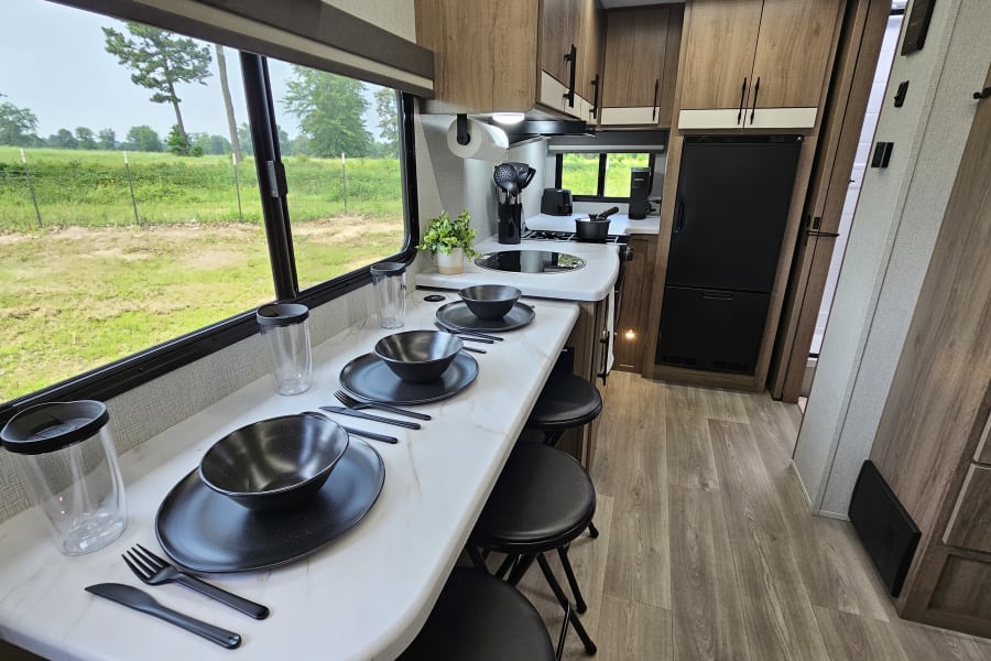 Clean RV rental kitchen and dining area featuring a modern countertop, bar stools, black place settings, and a scenic window.