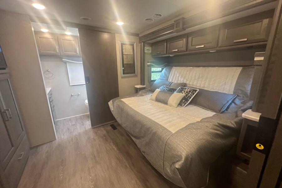 Spacious RV rental bedroom featuring a comfy bed, overhead cabinets, and an attached modern bathroom with wood-look flooring.