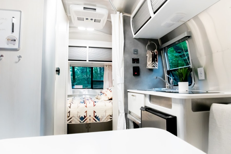 Cozy, modern RV rental interior with a compact kitchen, comfortable bedroom featuring a patterned quilt, and scenic forest views.