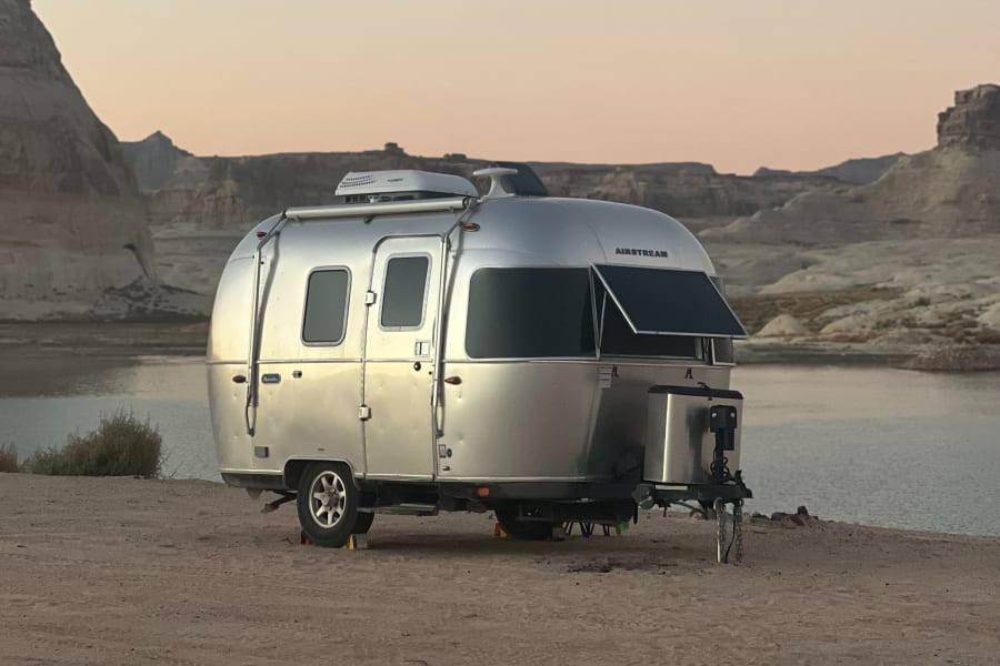 An Airstream RV rental camps by a serene lake at dusk, surrounded by rugged desert mountains, offering a perfect outdoor escape.