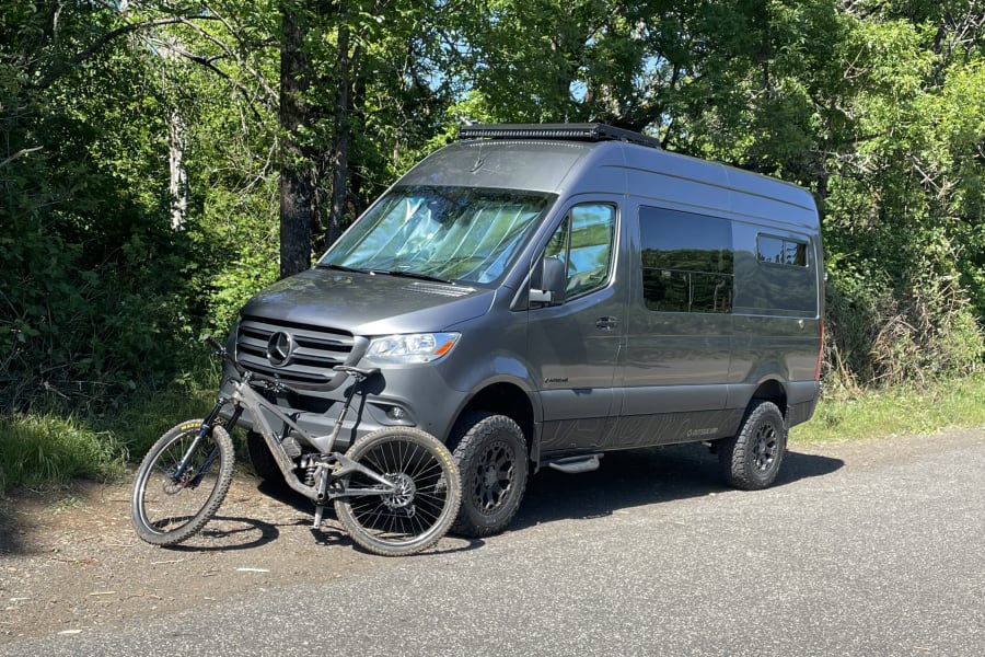 Gray Mercedes camper van (likely an **RV rental**), with a mountain bike, is parked on an asphalt road next to green trees, ready for adventure.