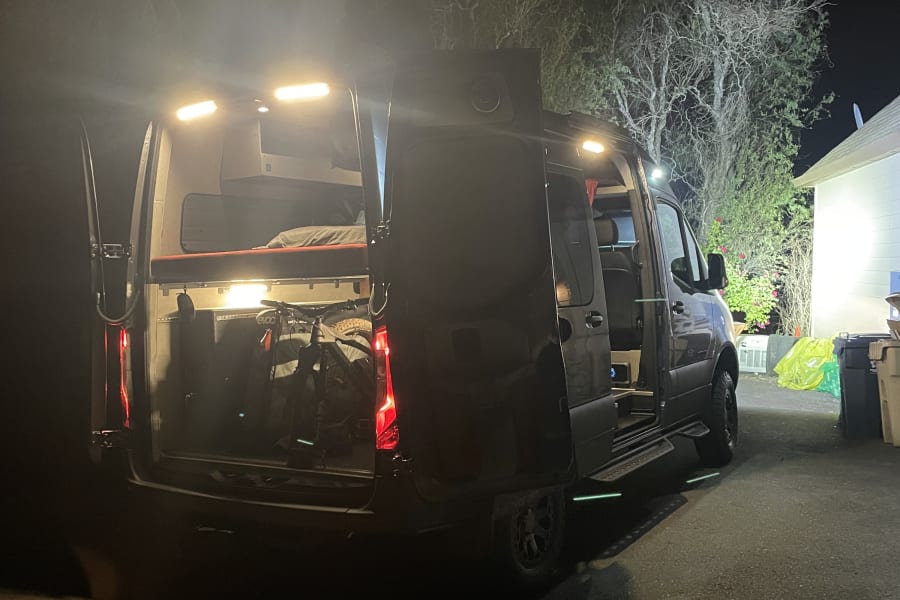 Illuminated RV rental at night with open rear and side doors, revealing a cozy bed and bike inside.