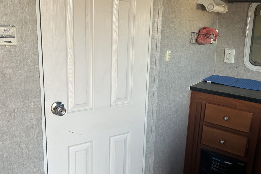 Inside an RV rental: features a white door, compact kitchen with brown cabinets & counter, plus a wall thermostat.