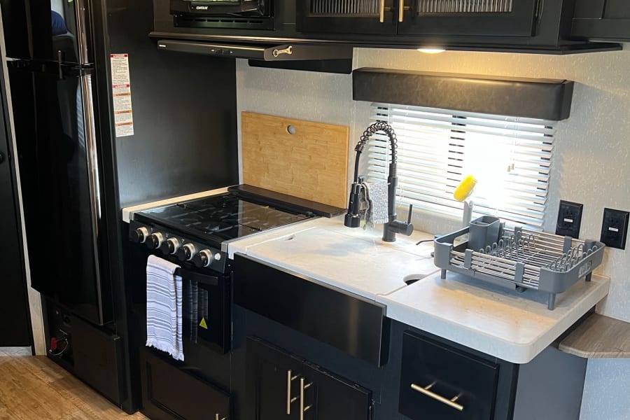 Modern RV rental kitchen: black cabinets, full-size fridge, stove, microwave, and large sink with white countertops.