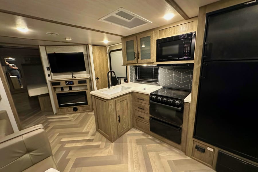 Modern RV rental interior featuring a compact kitchen, entertainment center with TV/fireplace, and comfortable seating.