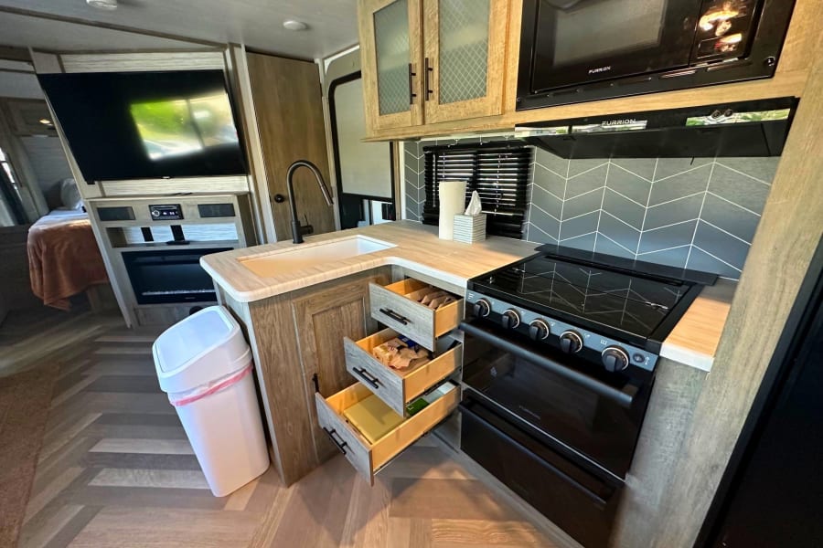 A well-equipped RV rental kitchen featuring a stove, microwave, sink, and multiple open storage drawers.