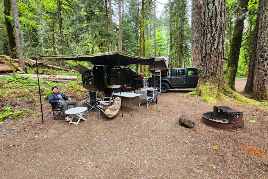 An RV rental overlanding adventure at a forest campsite. A man relaxes beside a rugged trailer & Jeep with a rooftop tent.
