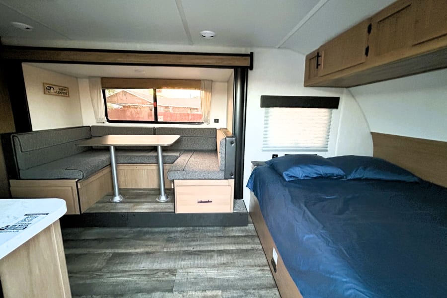Modern rv rental interior: comfortable dinette, cozy bed with blue sheets, wood-look floor, and overhead cabinets.