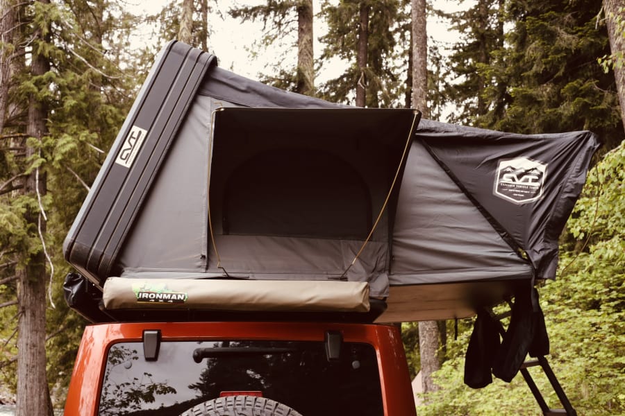 An open hardshell rooftop tent on a red SUV in a forest, providing a compact vehicle camping alternative to an RV rental.