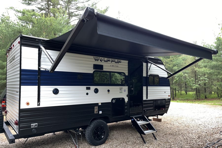Spacious Wolf Pup RV rental with extended awning parked on gravel at a wooded campsite, ready for adventure.