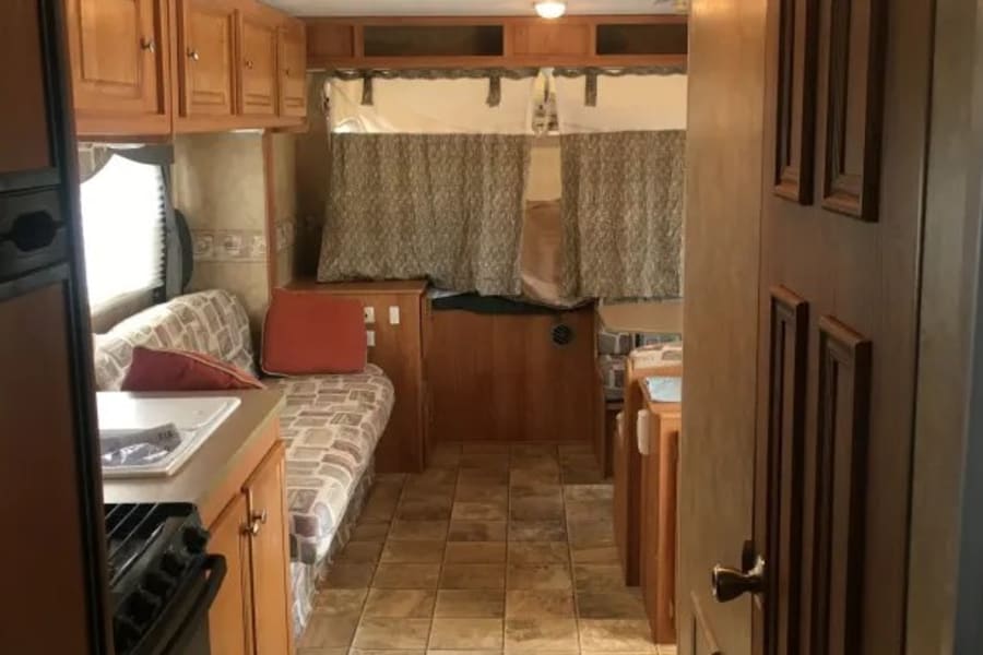 An RV rental interior view showing a kitchen, dinette with patterned cushions, and a potential sleeping area under curtains.