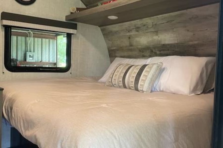 RV rental bedroom: a comfortable bed with pillows, overhead shelf, window, and wall clock. Cozy and well-appointed.