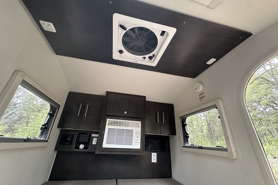 Inside a compact RV rental: featuring windows, overhead fan, AC, and dark wood cabinetry over the bed area.
