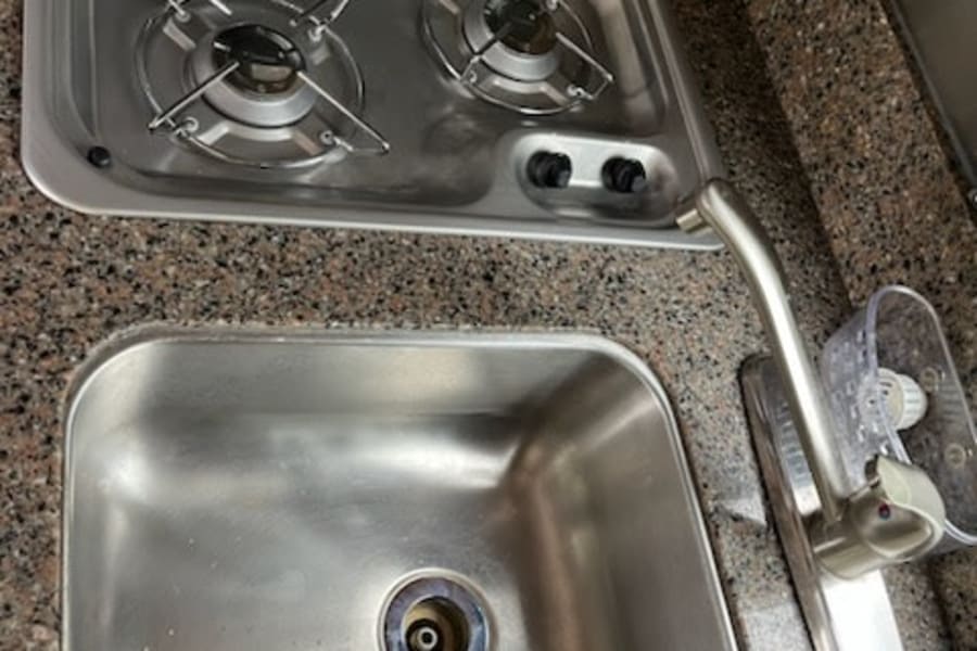 Compact RV rental kitchenette: two-burner stove, stainless steel sink, and faucet on a speckled countertop.