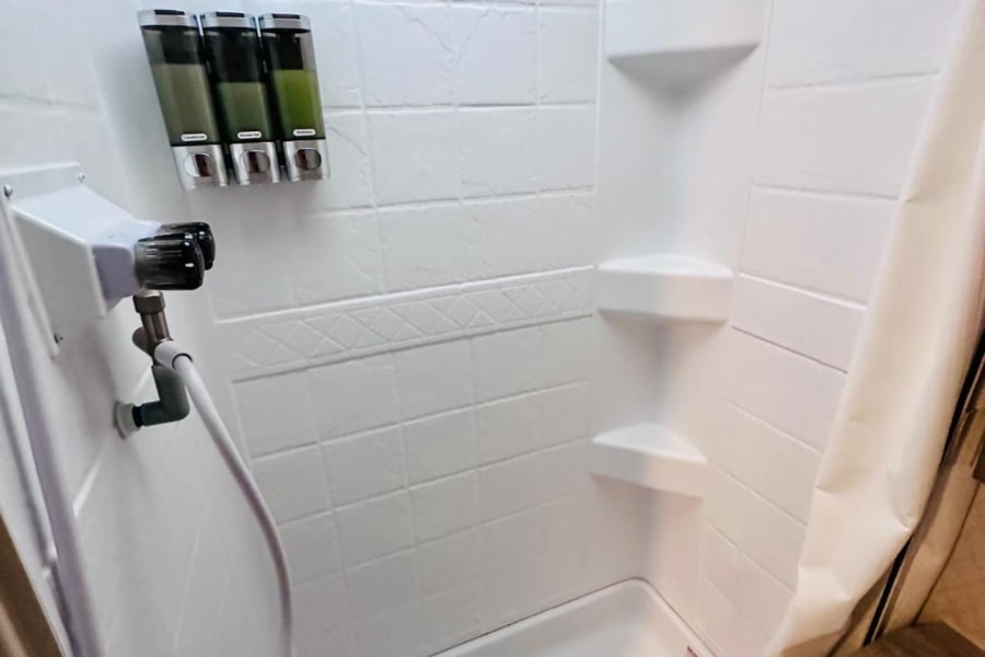 Clean, compact RV rental shower with wall-mounted soap dispenser, corner shelves, and an attached shower head.