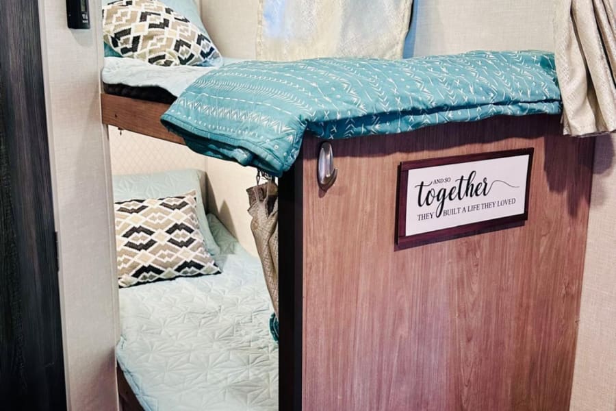 Cozy RV rental interior showing bunk beds with light blue bedding and patterned pillows. A welcoming sleep area.