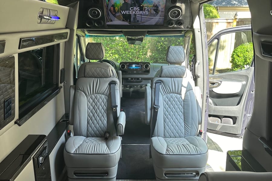 View inside a custom luxury RV rental with four light-gray captain's chairs, wood floor, and front TV screen.