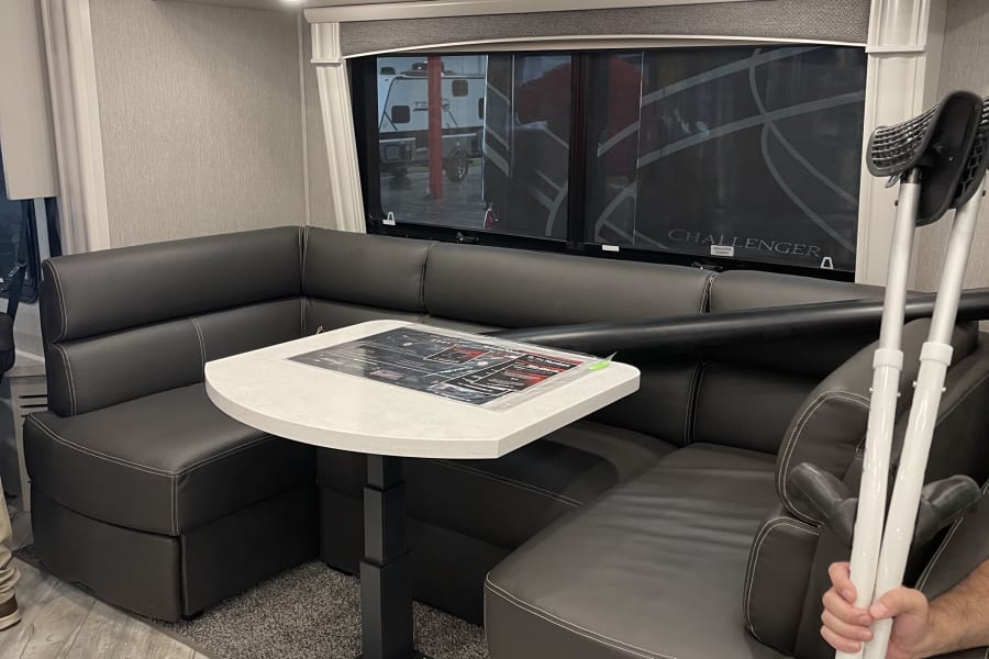 Inside an RV rental: L-shaped dinette with dark seating, white table, and overhead cabinets. Spacious layout for travel.