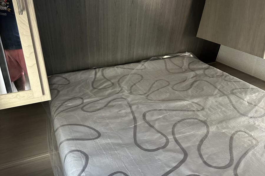 RV rental bed area: A new mattress with plastic wrap, sleek wood-grain interior, and overhead storage for travel.