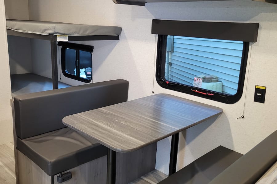 Inside this RV rental: a practical dinette with wood-grain table, dark grey seating, bunk beds, and overhead cabinets.
