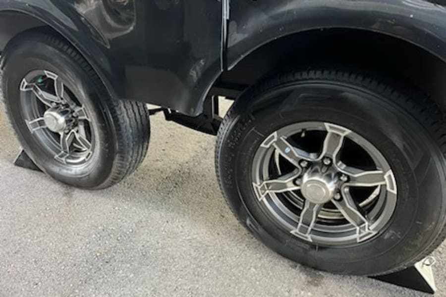 RV rental with tandem axles: close-up of two sets of black wheels, gray alloy rims, and tires parked on pavement, secured by chocks.