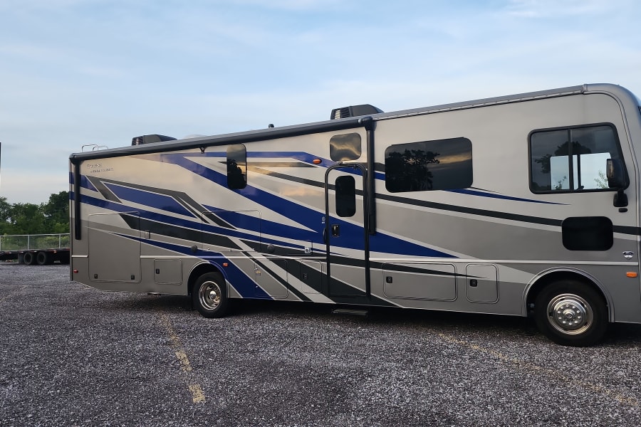 A large, silver and blue motorhome RV rental is parked on a gravel lot, waiting under a clear sky.