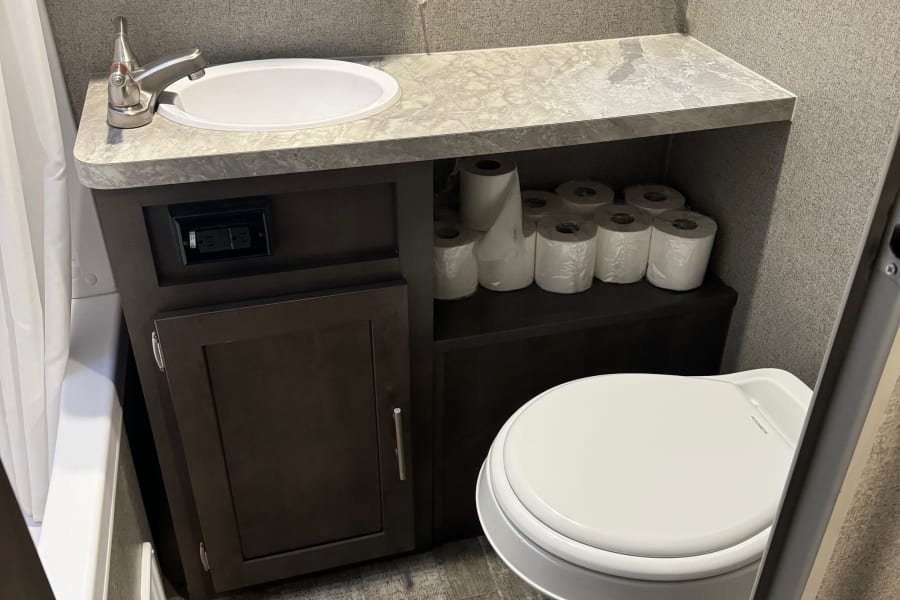 RV rental bathroom with a compact sink, toilet, and window. Organized storage for toilet paper. Ideal for your next road trip.